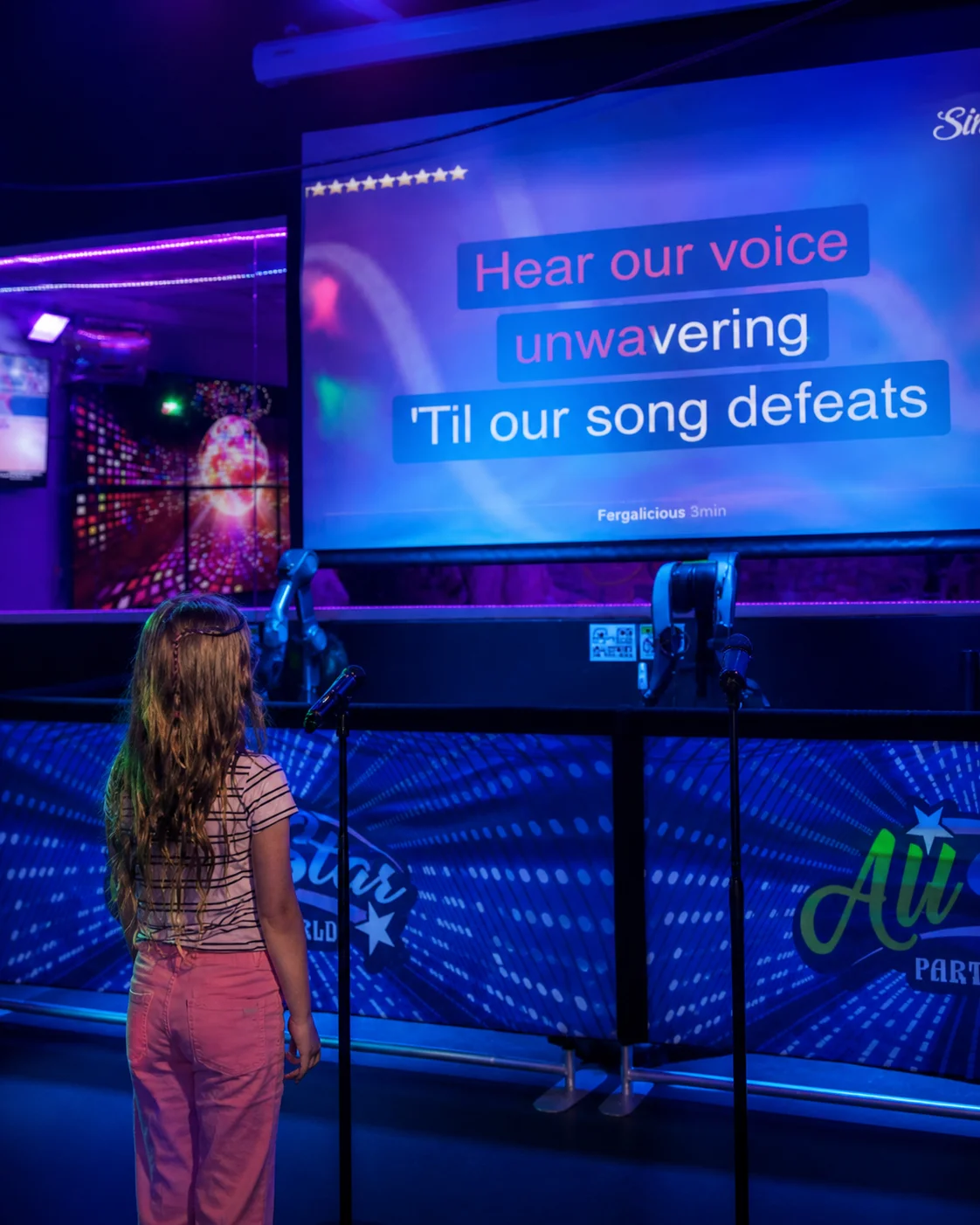 Young girl singing into a mic in front of a large projection screen at All Star Party World