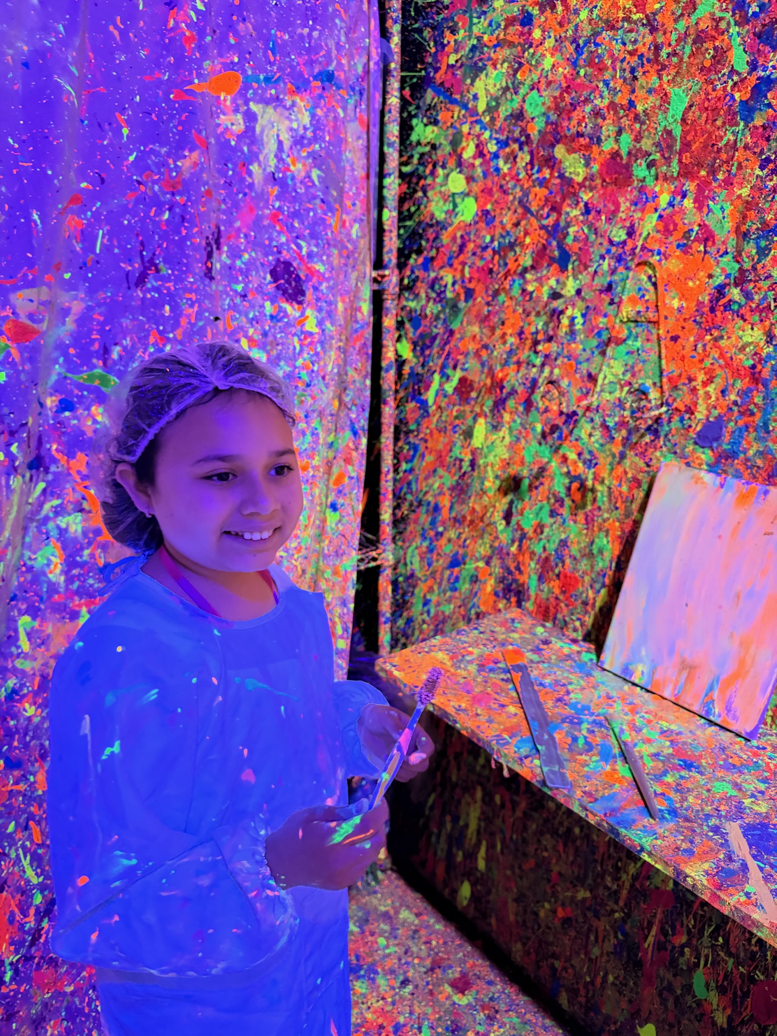 Girl smiling in white smock inside the neon blacklight Paint Me One room