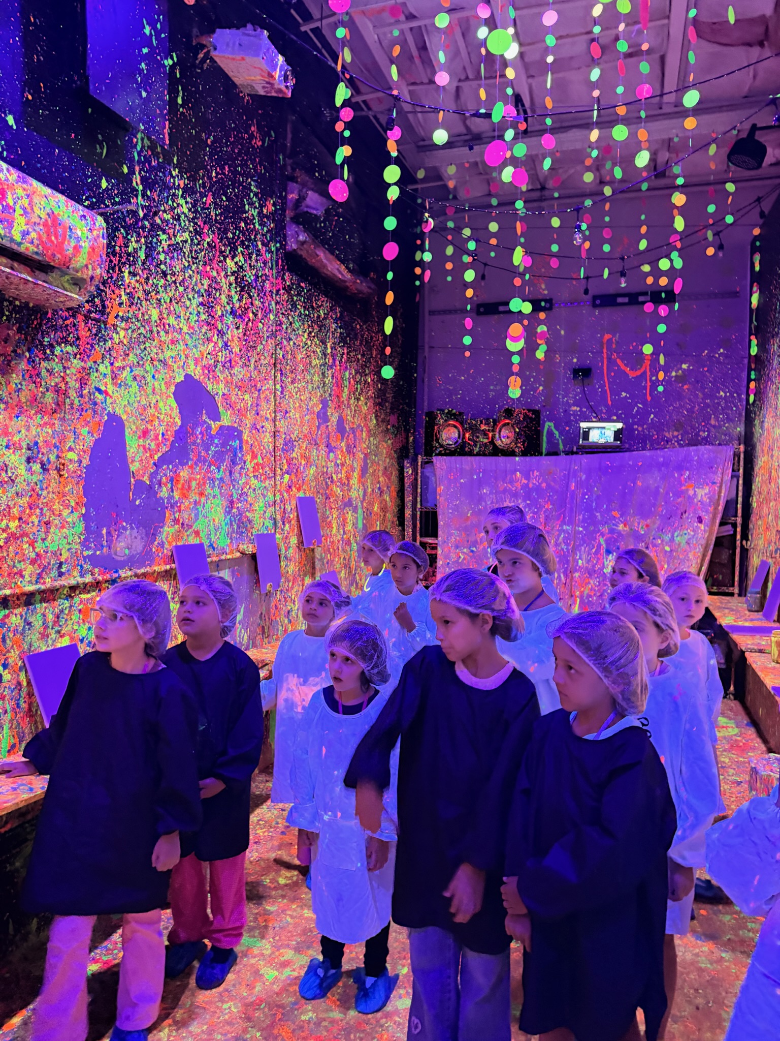 Large group of participants in white smocks standing in the neon paint-splattered blacklight room