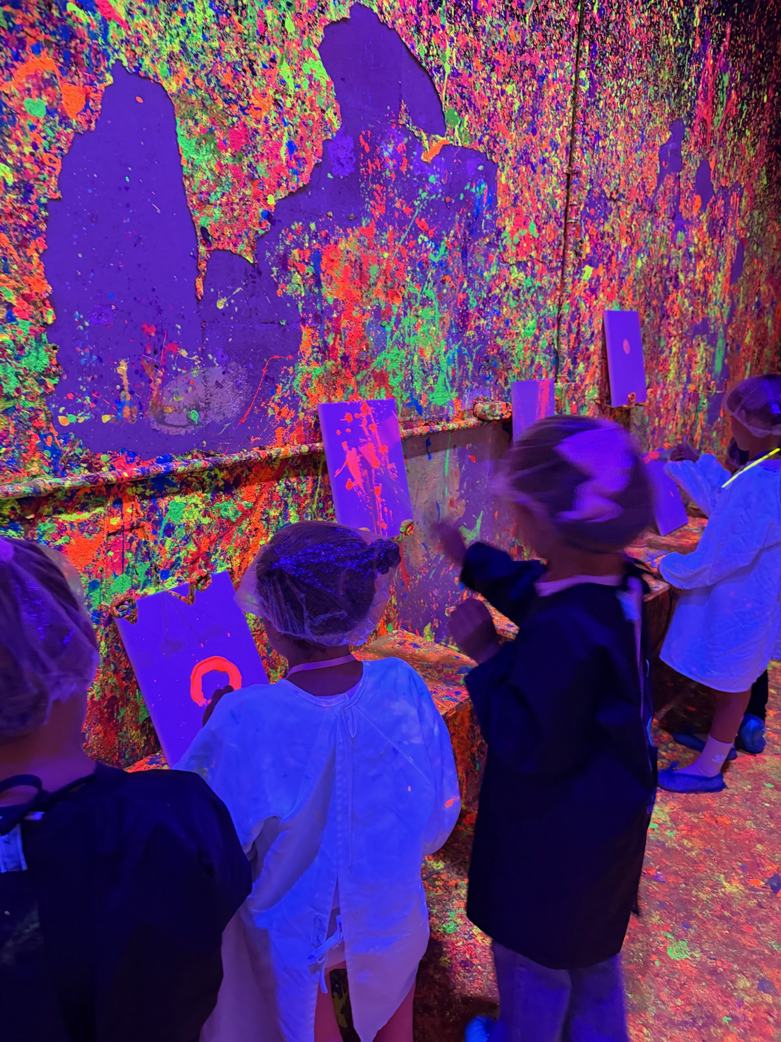 Kids painting on canvases in front of the neon splatter wall under UV blacklights