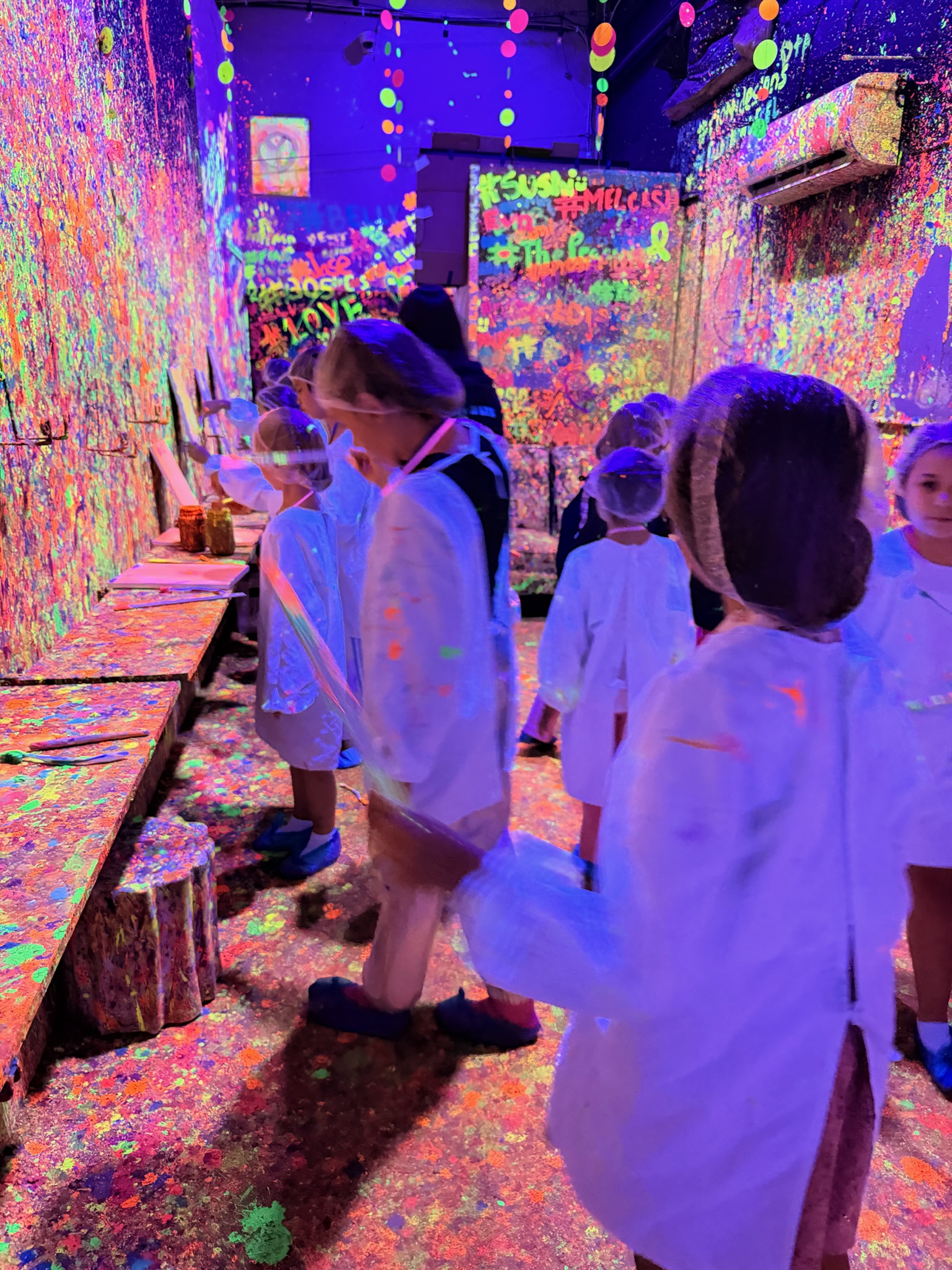Wide angle view of kids in white lab coats in the Paint Me One room with colorful hanging dot decorations